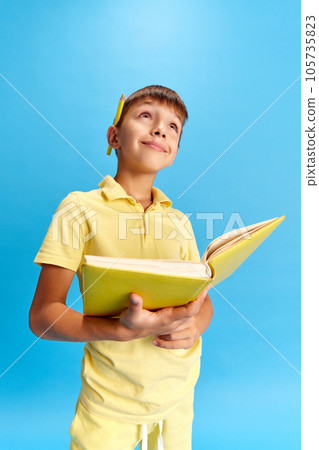 Emotive school boyholding book with dreamy and positive face against blue studio background Emotive school boyholding book with dreamy and positive face against blue studio background 105735823