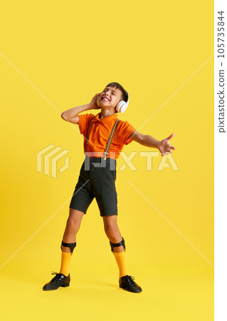 Full-length of little boy, child in stylish clothes listening to music in headphones against yellow studio background Full-length of little boy, child in stylish clothes listening to music in headphones against yellow studio background 105735844