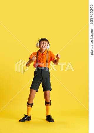 Full-length of little boy, child in stylish clothes listening to music in headphones against yellow studio background Full-length of little boy, child in stylish clothes listening to music in headphones against yellow studio background 105735846