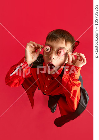 Top view. Little boy, child in costume for halloween party holding fake eyes on glasses on face against red studio background Top view. Little boy, child in costume for halloween party holding fake eyes on glasses on face against red studio background 105735855
