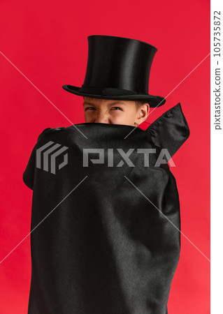 Emotive little boy, child in hat covering face with black cloak against red studio background. Costume party Emotive little boy, child in hat covering face with black cloak against red studio background. Costume party 105735872