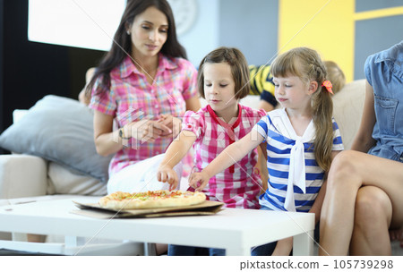 Little girls with mothers sit on couch and take slices of pizza Little girls with mothers sit on couch and take slices of pizza 105739298