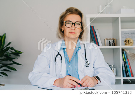 Portrait of confident mature middle aged woman doctor medical nurse portrait, female physician practitioner in white coat with stethoscope sitting at her working place at hospital office. 105741353