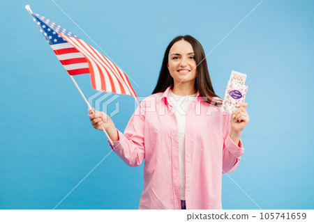 Young brunette woman in pink shirt poses on blue background in studio with american flag and hundred dollar cash with passport. Concept of success, travel, knowledge of foreign language 105741659
