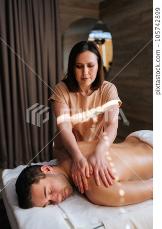Vertical portrait of focused female masseuse doing shoulder massage to relaxed man in spa center lying closed eyes. Woman massage therapist performing circular movements to relax back muscles. 105742899