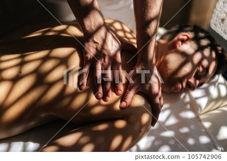 Close-up hands of unrecognizable masseur male doing back spa massage of man in spa massage room lying on table in sunlight and shadows. Relaxed client came to massage therapy due to body aches. Close-up hands of unrecognizable masseur male doing back spa massage of man in spa massage room lying on table in sunlight and shadows. Relaxed client came to massage therapy due to body aches. 105742906