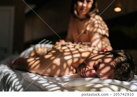 Selective focus cropped shot of female masseuse giving massage to handsome man in luxury spa salon lying closed eyes on massage table in sunlight and beautiful shadows. Concept of healthy lifestyle. Selective focus cropped shot of female masseuse giving massage to handsome man in luxury spa salon lying closed eyes on massage table in sunlight and beautiful shadows. Concept of healthy lifestyle. 105742910