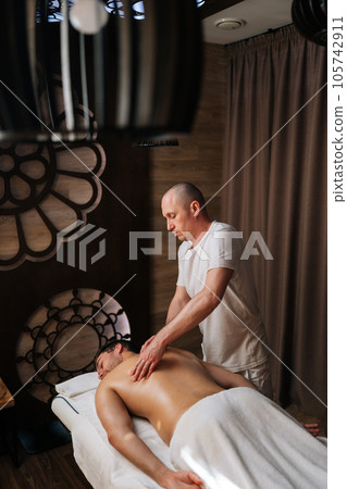Remote rear view of professional male massage therapist using massage oil for deep muscle relaxation in spa center. Relaxed young man enjoying being gently massaged lying on table with closed eyes Remote rear view of professional male massage therapist using massage oil for deep muscle relaxation in spa center. Relaxed young man enjoying being gently massaged lying on table with closed eyes 105742911