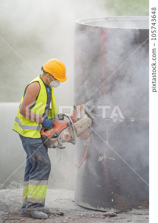 Installing a concrete pipe in the ground to collect rainwater. The worker cuts holes in the concrete part of the pipe to connect to the storm drain. A man in construction clothes is sawing concrete. Installing a concrete pipe in the ground to collect rainwater. The worker cuts holes in the concrete part of the pipe to connect to the storm drain. A man in construction clothes is sawing concrete. 105743648
