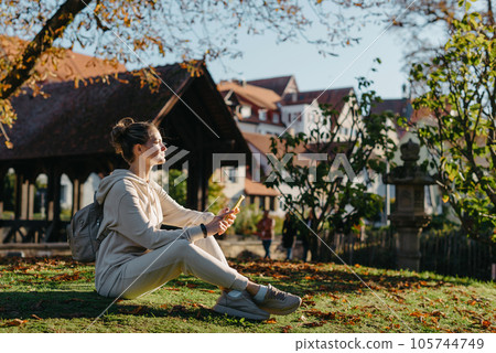 Young Fashionable Teenage Girl With Smartphone In Europian Park In Autumn Sitting At Smiling. Trendy Young Woman In Fall In Park Texting. Retouched, Vibrant Colors. Beautiful Blonde Teenage Girl 105744749