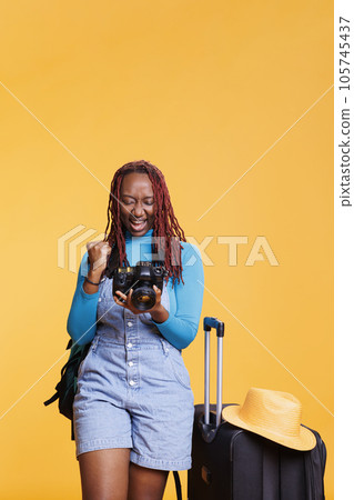 Cheerful adult checking pictures on dslr, looking at photos of architecture buildings and landscapes in studio with suitcase. Woman sightseeing and taking photographs on holiday. 105745437