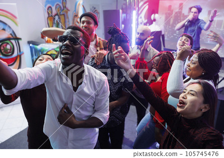 Happy friends group posing for selfie together while having fun and relaxing at nightclub party. Cheerful young diverse people crowd screaming and laughing while taking photo on dancefloor 105745476