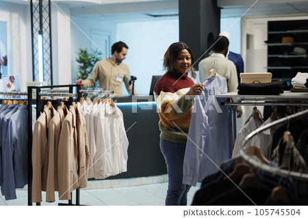 Woman holding shirt on rack and looking at camera while choosing outfit in clothing store. Smiling african american customer selecting formal wear and shopping in boutique portrait 105745504