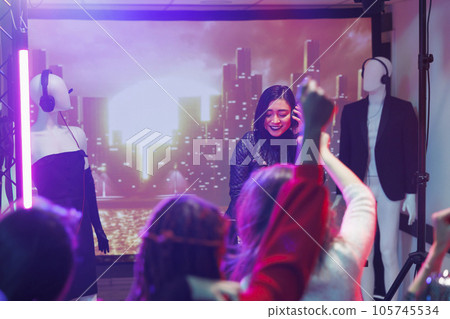 People attending techno music concert in nightclub while asian dj performing. Woman musician holding headphones while mixing sound using controller during party in nightclub 105745534