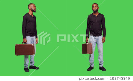 Casual person holding suitcase luggage on camera and waiting for something, standing over green screen backdrop. Young man with briefcase carrying baggage in studio, friendly adult. 105745549