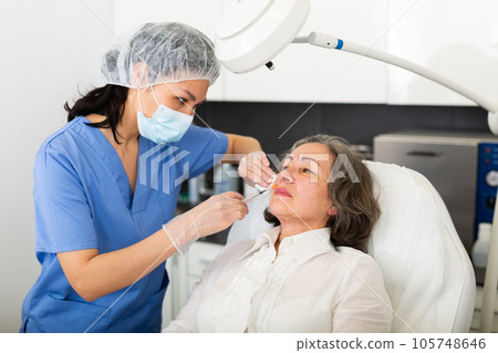 Young cosmetologist doctor in a protective mask makes an elderly woman patient a injecting an injection into the face 105748646