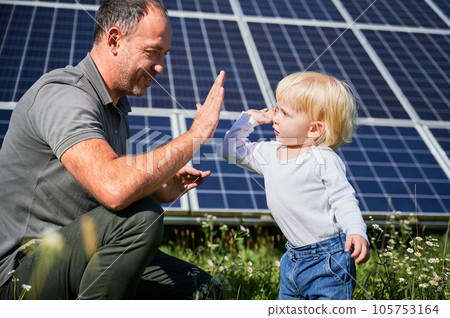 Smiling father and his little child giving each other high-five on background of solar panels. Young father enjoy spending time with his son. Happy family of two on background of solar panels. 105753164