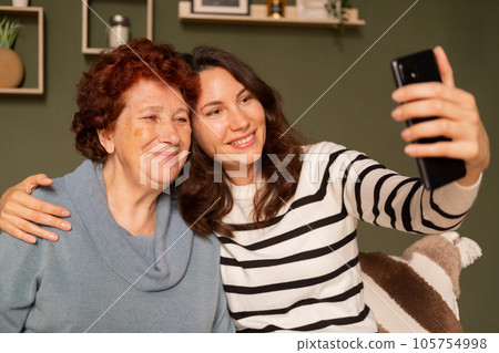 Portrait of a modern happy grandmother and granddaughter sitting on the sofa during the day in the living room. A young woman hugs an elderly woman by the shoulder and they take a selfie with a smile 105754998