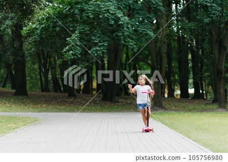 Active little child girl riding scooter on road in park outdoors on summer day Active little child girl riding scooter on road in park outdoors on summer day 105756980