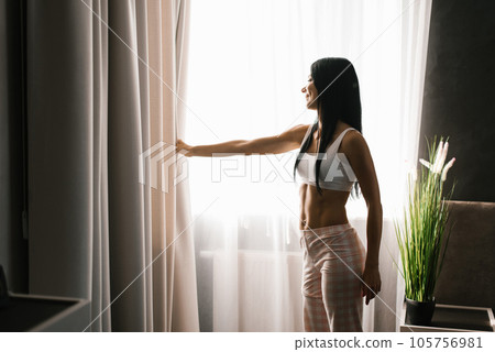 Brunette woman stands at the bedroom window in the morning. She has parted the curtains on the window and is looking at the street 105756981