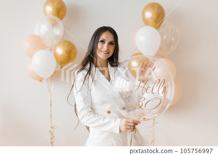 Positive woman celebrating anniversary dressed in stylish feather dress posing with birthday balloon around inflated balloons at home 105756997