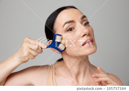 Mature woman using facial roller for smooth, anti aging and lifting treatment on a grey studio background. 105757384