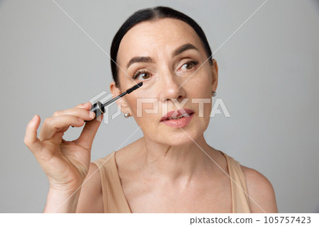 Close up portrait of beautiful mature brunette woman applying mascara with makeup brush over grey background. Fashion beauty. Close up portrait of beautiful mature brunette woman applying mascara with makeup brush over grey background. Fashion beauty. 105757423
