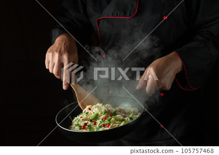 The chef fries spaghetti with spices and seasonings in a hot pan with steam. Space for advertising on a black background. Italian national dish. The chef fries spaghetti with spices and seasonings in a hot pan with steam. Space for advertising on a black background. Italian national dish. 105757468