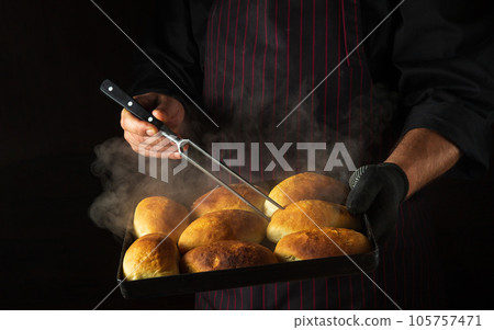 The baker bakes pies or buns on a hot baking sheet taken out of the oven. Checking readiness with a fork in the chef hand. Black space for menu or recipe 105757471