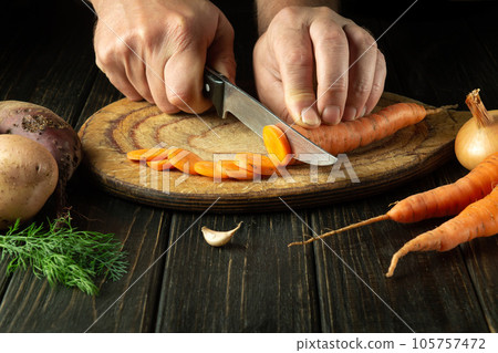 The cook cuts fresh carot on a wooden cutting board. Close-up of chef hands while preparing vegetarian food 105757472