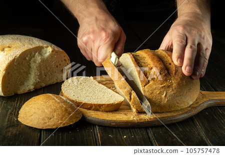 The chef cuts wheat bread on the wooden board of the bakery kitchen for tasting. Healthy food concept on black background The chef cuts wheat bread on the wooden board of the bakery kitchen for tasting. Healthy food concept on black background 105757478