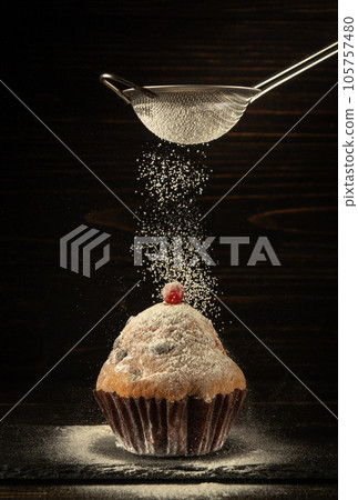 From the sieve, powdered sugar is sprinkled onto a baked red berry muffin. Delicious dessert concept on black background. From the sieve, powdered sugar is sprinkled onto a baked red berry muffin. Delicious dessert concept on black background. 105757480