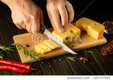 The hands of the chef with a knife cut the cheese into small pieces for tasting. Delicious milk cheese recipe presentation with rosemary and pepper 105757485