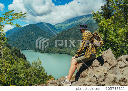 Hiking traveller caucasian bearded man with backpack sitting on rocks enjoying beautiful view of turquois blue Lake Ritsa in Abkhazia 105757811