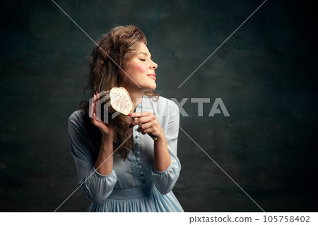 Portrait charming aristocratic woman wearing blue historical dress holding vintage hairbrush in hand and brushing her hair. Portrait charming aristocratic woman wearing blue historical dress holding vintage hairbrush in hand and brushing her hair. 105758402