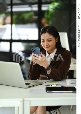 Charming asian woman entrepreneur sitting in bright home office and using smartphone. Business, technology and people Charming asian woman entrepreneur sitting in bright home office and using smartphone. Business, technology and people 105758589