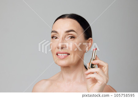 Beautiful old age. Photo of elderly well-kept brunette woman holding lipstick over grey studio background. Make up. Beautiful old age. Photo of elderly well-kept brunette woman holding lipstick over grey studio background. Make up. 105758595