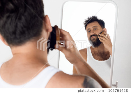 Mature man looking in mirror, taking care after beard, brushing with special brush against white studio background. Mature man looking in mirror, taking care after beard, brushing with special brush against white studio background. 105761428