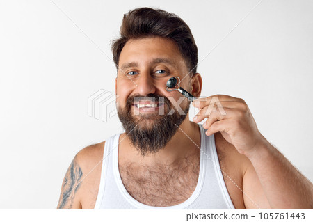 Smiling, bearded man doing face massage with roller against white studio background. Removing puffiness 105761443