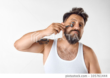 Morning refreshment. Bearded man taking care after skin, doing face massage with roller against white studio background 105761444