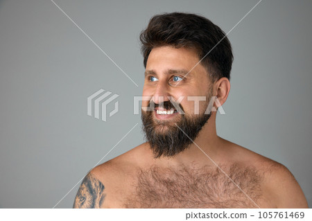 Handsome, smiling, mature, bearded man with blue eyes standing shirtless, looking away against grey studio background Handsome, smiling, mature, bearded man with blue eyes standing shirtless, looking away against grey studio background 105761469