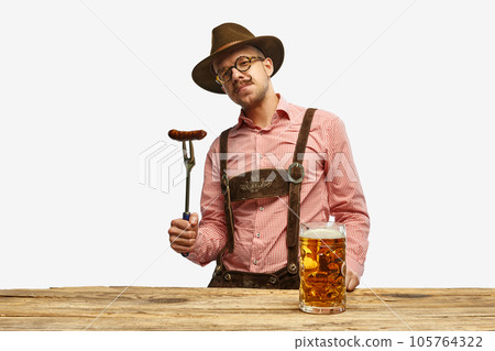 Bavarian mustache man in hat wearing traditional fest outfit holding hot fried sausage and looking at huge glass of beer. Oktoberfest concept Bavarian mustache man in hat wearing traditional fest outfit holding hot fried sausage and looking at huge glass of beer. Oktoberfest concept 105764322