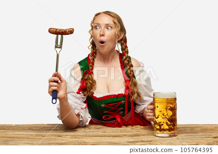 Surprised young woman dressed traditional dirndl, holding huge juicy,appertizing Bavarian sousage. Concept of Oktoberfest. 105764395