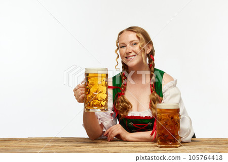 Smiling young girl waitress, wearing traditional Bavarian or german dirndl, serving huge beerglasses over white background. Concept of Oktoberfest. 105764418