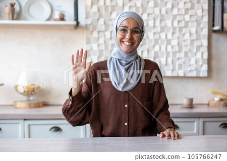 Muslim young woman in hijab sitting in the kitchen at home and smiling talking and greeting to the camera. 105766247