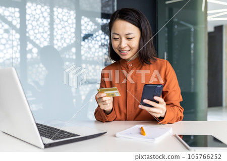 Young beautiful and successful Asian woman at workplace inside office, businesswoman holding phone and bank credit card in hands, female employee making online purchases using smartphone app. 105766252