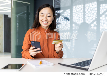 Young beautiful and successful Asian woman at workplace inside office, businesswoman holding phone and bank credit card in hands, female employee making online purchases using smartphone app. 105766253