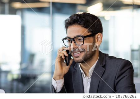 Young successful arab businessman boss talking on phone close up, man smiling contentedly inside office at workplace in business suit. Young successful arab businessman boss talking on phone close up, man smiling contentedly inside office at workplace in business suit. 105766262