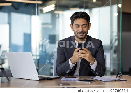 Successful experienced businessman using phone while sitting at workplace, hispanic smiling happy with achievement results holding phone, reading online using app on smartphone. 105766264