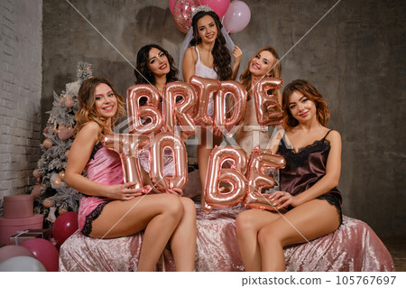 Ladies in sexy lingerie and bride in veil, enjoying a hen-party, sitting on bed, holding balloons, smiling. New year tree, decorations. Close-up. 105767697